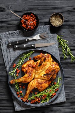 butterflied or spatchcock roast whole chicken on dark grey plate with rosemary and cranberry on black wooden table with cutlery and ingredients, vertical view from above clipart