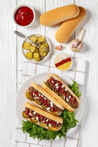 stock image Midwestern Loose Meat Sandwiches, Juicy burgers of ground beef topped with pickles, raw onion and ketchup on plate on white wooden table with ingredients, vertical view from above
