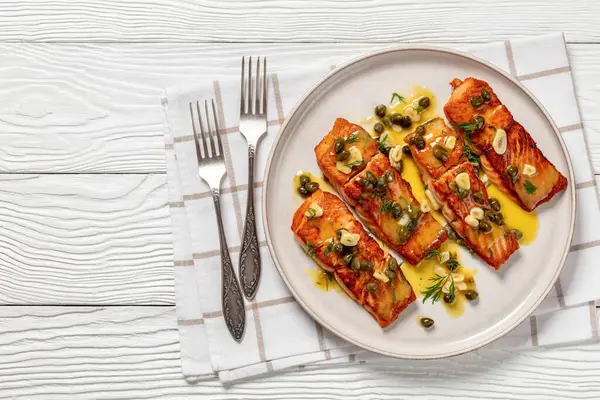 Stock image salmon piccata, fish fillet with crispy golden crust with tangy lemon caper sauce on a plate on white wooden table with forks, horizontal view from above, flat lay, free space