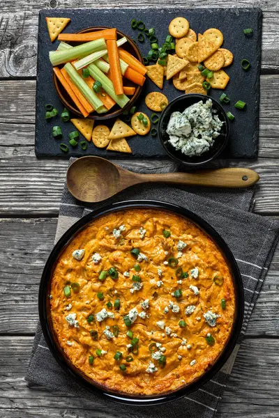 stock image buffalo chicken dip in baking dish on grey rustic wooden table with crackers, blue cheese, carrot and celery sticks on slate board, vertical view from above