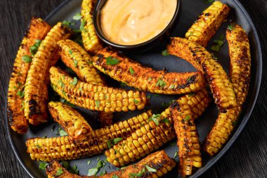 bbq corn ribs, seasoned juicy corn kernels sprinkled with fresh parsley on plate with paprika mayo dipping sauce on plate on dark wooden table, close-up, dutch angle view clipart