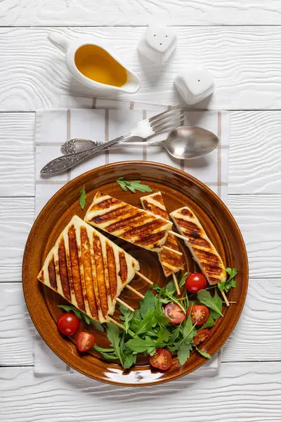 stock image Grilled Greek Halloumi Cheese on sticks with fresh rocket salad, tomatoes on brown plate on white wooden table with fork and spoon, honey olive oil sauce, vertical view