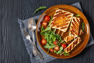 Grilled Halloumi Cheese on sticks with fresh rocket salad, tomatoes on plate on black wooden table with fork and spoon, horizontal view from above, flat lay, free space clipart