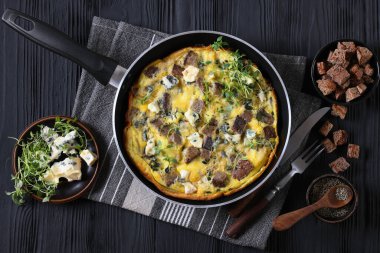 simple savory omelette with blue cheese, rye croutons and fresh thyme on a skillet on black wooden table with cutlery and ingredients, horizontal view from above, flat lay, close-up clipart