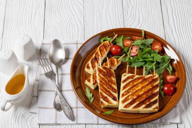 Grilled Greek Halloumi Cheese on sticks with fresh rocket salad, tomatoes on brown plate on white wooden table with fork and spoon, honey olive oil sauce, close-up, horizontal view clipart