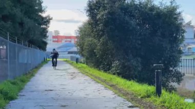 Sanayi manzarası ve doğal yeşilliklerle çevrili Petaluma Nehri 'nin kıyısındaki yolda çalışmak için bisiklet süren bir adam.