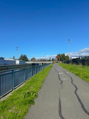 Açık mavi bir gökyüzünün altında, uzak kırmızı bir heykele doğru uzanan, çimenli kenarlı, asfalt bir yolun güneşli gündüz manzarası