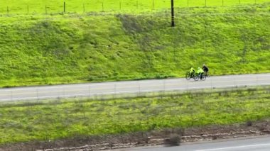Parlak giyinmiş bir bisikletçi, canlı yeşil bir yamaçtan geçen yol boyunca pedal çeviriyor. Aktif kırsal yaşamın bir resmi.
