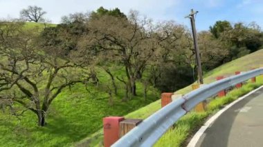 Yol kenarı manzarası. Parmaklıkları ve trafik levhası var. Arkaplanı yemyeşil tepeler ve uzak bir otoyol.