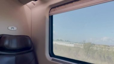 Bir tren penceresinden içeriden bakıldığında, açık araziler ve açık mavi gökyüzünün altındaki uzak binalarla kırsal kesimin kısa bir görüntüsünü gösteriyor.