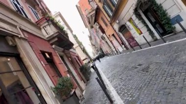 Roma 'da kaldırım taşı yolu boyunca binalar ve sokak lambaları olan eğimli bir cadde manzarası. Kamera döndürme efekti şehir sahnesine bir hareket ve benzersizlik hissi ekler..
