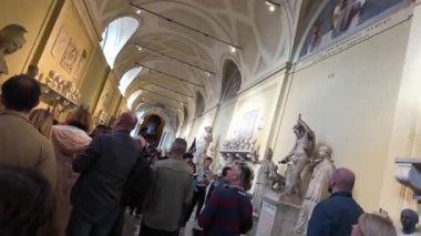Turistler Vatikan Müzesi 'nin zengin dekore edilmiş bir koridorunu klasik büstler ve başka bir galeriye uzanan ayrıntılı kemerlerle dolduruyorlar. Sahne, sanat ve mimarinin karışımını yakalar..