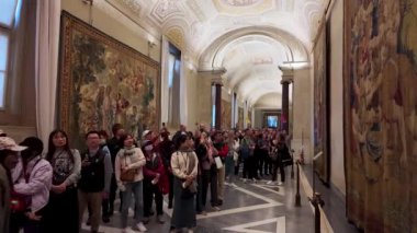 Turistler Vatikan Müzesi 'nin duvar halısı galerisinden geçerek duvarlarda asılı olan karmaşık duvar halılarını izliyorlar. Galerinin kubbeli tavanı ve detaylı dekor ihtişama katkı sağlıyor..