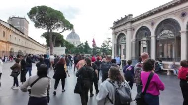 Turist kalabalığı Vatikan Müzeleri 'nin avlusunu keşfederken, arka planda Aziz Peter Bazilikası görülüyor. Sahne, ziyaretçilerin tarihi manzaranın tadını çıkarmasıyla dolu..