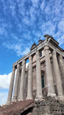 Roma 'daki Satürn Tapınağı' nın çarpıcı dikey görüntüsü, yüksek antik sütunlarını dağınık bulutlu parlak mavi gökyüzüne karşı sergiliyor. Bu resim, bu ikonik Roma yapısının ihtişamını ve tarihsel önemini yakalar..