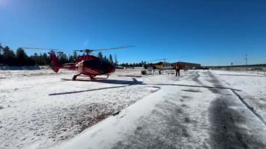 Büyük Kanyon yakınlarındaki karlı bir pistte, açık mavi bir gökyüzünün altında birkaç helikopter park halindedir. Sahne, kış aylarında uçmadan önceki hazırlık ve sükuneti yakalar..