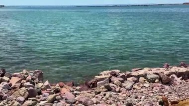 Parlak bir gökyüzünün altında ufka uzanan kristal berrak mavi suyu olan kayalık bir sahil manzarası. Huzur ve doğa temaları için mükemmel..