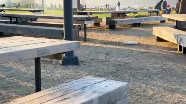 Açık çakıl pikniği alanı, ahşap banklar ve uzak insanlarla, akşam güneşiyle yıkanmış ve huzurlu atmosferle.