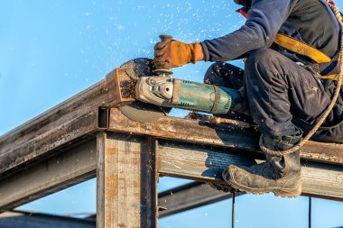 İşçi inşaat alanında çelik kutu profilini el değirmeni makinesiyle öğütüyor. Öğütme, kesme aracı olarak öğütme tekeri kullanan yıpratıcı bir makine işlemidir.