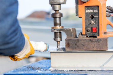 Worker is making a hole in the steel structure material with portable magnetic drill machine in the construction site. It can use twist drill bits, annular cutters and milling cutters.