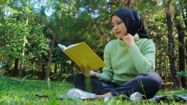 Rahat Müslüman kadın parkta eğleniyor, çimenlerde oturuyor ve kitap okuyor, boş bir alan.