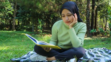 Rahat Müslüman kadın parkta eğleniyor, çimenlerde oturuyor ve kitap okuyor, boş bir alan.