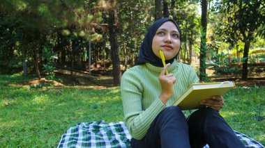 Parkta tesettürlü gülümseyen müslüman kadın elinde kitap ve kalem tutuyordu.