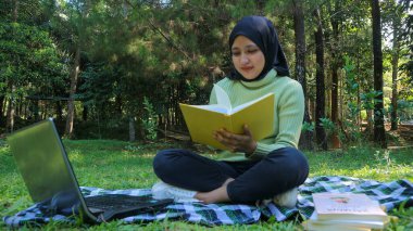 Rahat Müslüman kadın parkta eğleniyor, çimenlerde oturuyor ve kitap okuyor, boş bir alan.