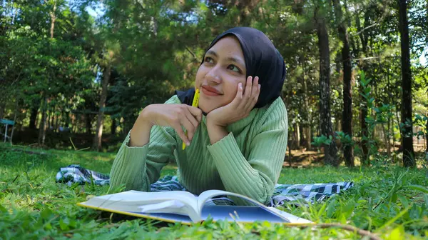 Smiling muslim woman in hijab in park holding book and pen thinking idea.Empty space