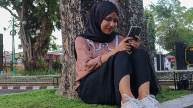 Şehir parkında cep telefonu kullanan, okuyan veya mesajları kontrol eden Müslüman bir kadının portresi. Keyifli, rahatlatıcı bir hafta sonu.