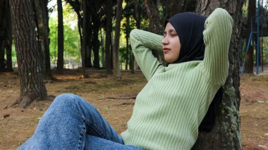 Elleri başının arkasında, gözleri kapalı, ağaca yaslanmış gülümseyen Müslüman kadın.