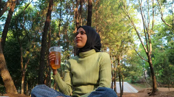 Çam ormanında elinde fincanla oturan başörtüsü takan Müslüman bir kadın portresi. Yaz güneşli parktaki modern Müslüman kadın. Kendine güvenen İslamcı kız.