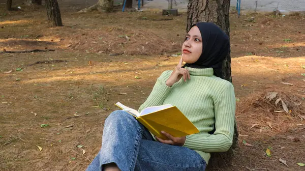 Genç Müslüman kadın başörtüsü takmış, parkta bir ağaca yaslanırken kitap okuyor.