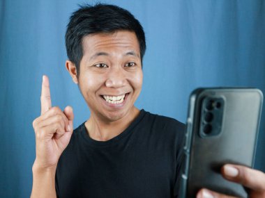 Komik gülümseyen yüz ifadesi. Asyalı adam gülümsüyor. Elinde cep telefonu var.