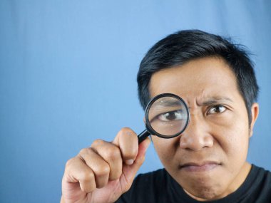 Komik ifadeli Asyalı genç adam büyüteçten bakıyor, bir şeyi arıyor ya da araştırıyor.