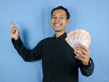 Heyecanlı bir yüz ifadesi. Yakışıklı Asyalı bir adam, bir kenara işaret ederek kağıt para banknotları tutuyor. İş ve finans kavramı.
