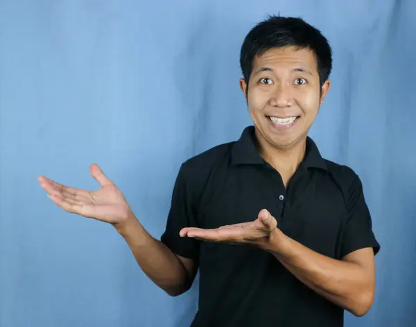 Portrait of young Asian man smiling with open palm gesture isolated on studio blue background for advertising and product placement concept