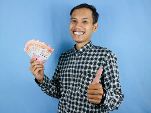 Heyecanlı bir yüz ifadesi. Yakışıklı Asyalı adam, elinde kağıt para banknotları ve iyi niyetli bir jest tutuyor. İş ve finans kavramı.