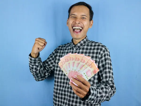 Heyecanlı yüz ifadesi, yakışıklı Asyalı adam, kağıt para banknotlarını yumruğunu sıkarak tutuyor. İş ve finans kavramı.