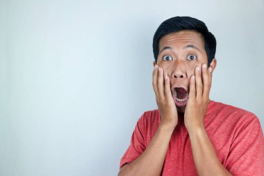 Kırmızı t-shirt giyen Asyalı adamın kameraya bakarken şaşkın ve şaşırmış ifadesi.