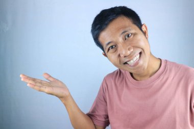 Gülen Asyalı genç adamın, sağ elini göstererek beyaz arka planda izole edilmiş ürünleri tanıttığı komik yüz ifadesi.