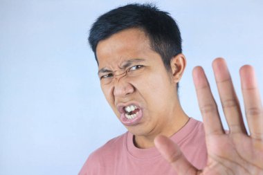 Komik surat ifadesi genç Asyalı adam kameraya bakarken el hareketi reddediyor, beyaz arka planda izole edilmiş.