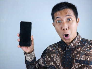 Şok olmuş Asyalı adam batik kıyafetler giyiyor. Akıllı telefonu boş ekran ile gösteriyor.