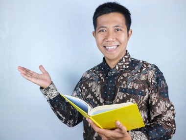 Batik kıyafetler giymiş gülümseyen Asyalı bir adam fotokopi odasını sunuyor ya da elinde kitap tutarken bir şeyler gösteriyor.