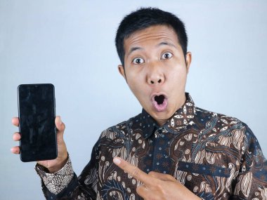 Şok olmuş Asyalı adam batik kıyafetler giyiyor. Akıllı telefonu boş ekran ile gösteriyor.