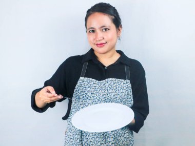 Önlüklü güzel Asyalı kadın beyaz arka planda boş beyaz tabak ya da tencere tutuyor.