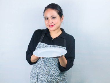 Önlüklü güzel Asyalı kadın beyaz arka planda boş beyaz tabak ya da tencere tutuyor.