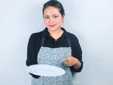 Önlüklü güzel Asyalı kadın beyaz arka planda boş beyaz tabak ya da tencere tutuyor.