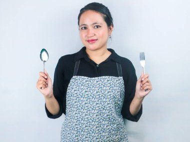 Önlüklü, elinde kaşıkla çatalla beyaz arka planda duran güzel Asyalı kadın.