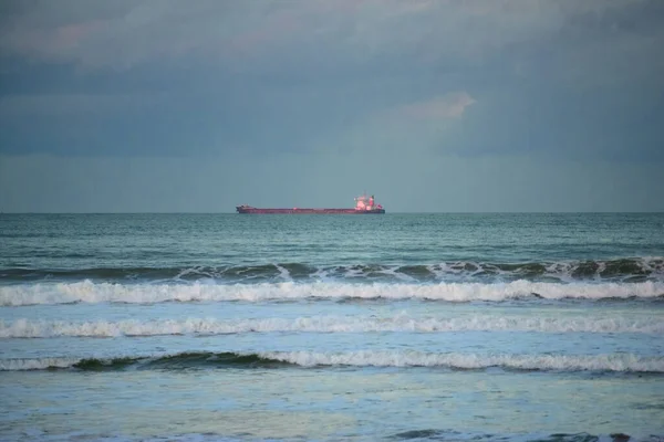 A cargo ship over the horizon under grey blue sky and sea with waves breaking on the near beach.